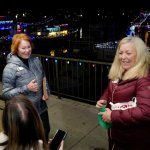 Two women smile outdoors at a colorful nighttime event with decorated boats.