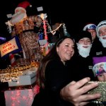 People take a selfie in festive hats near a Santa Claus decoration with lights at night.