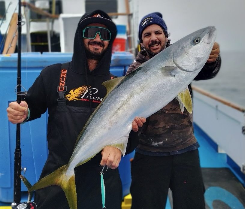 Two men on a boat holding a large fish, both smiling.