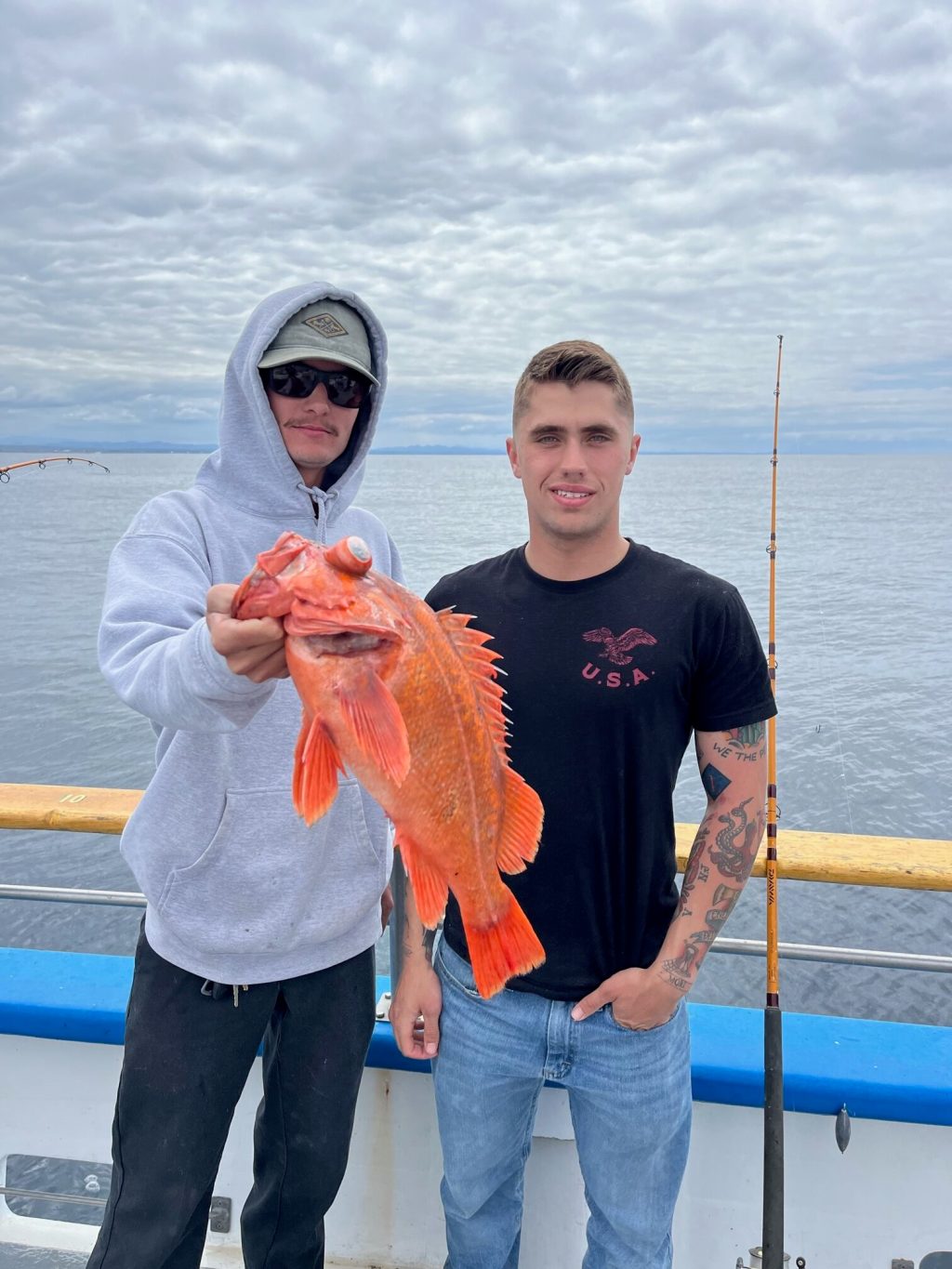 Two men on a boat, one holding a large red fish, cloudy sky in background.