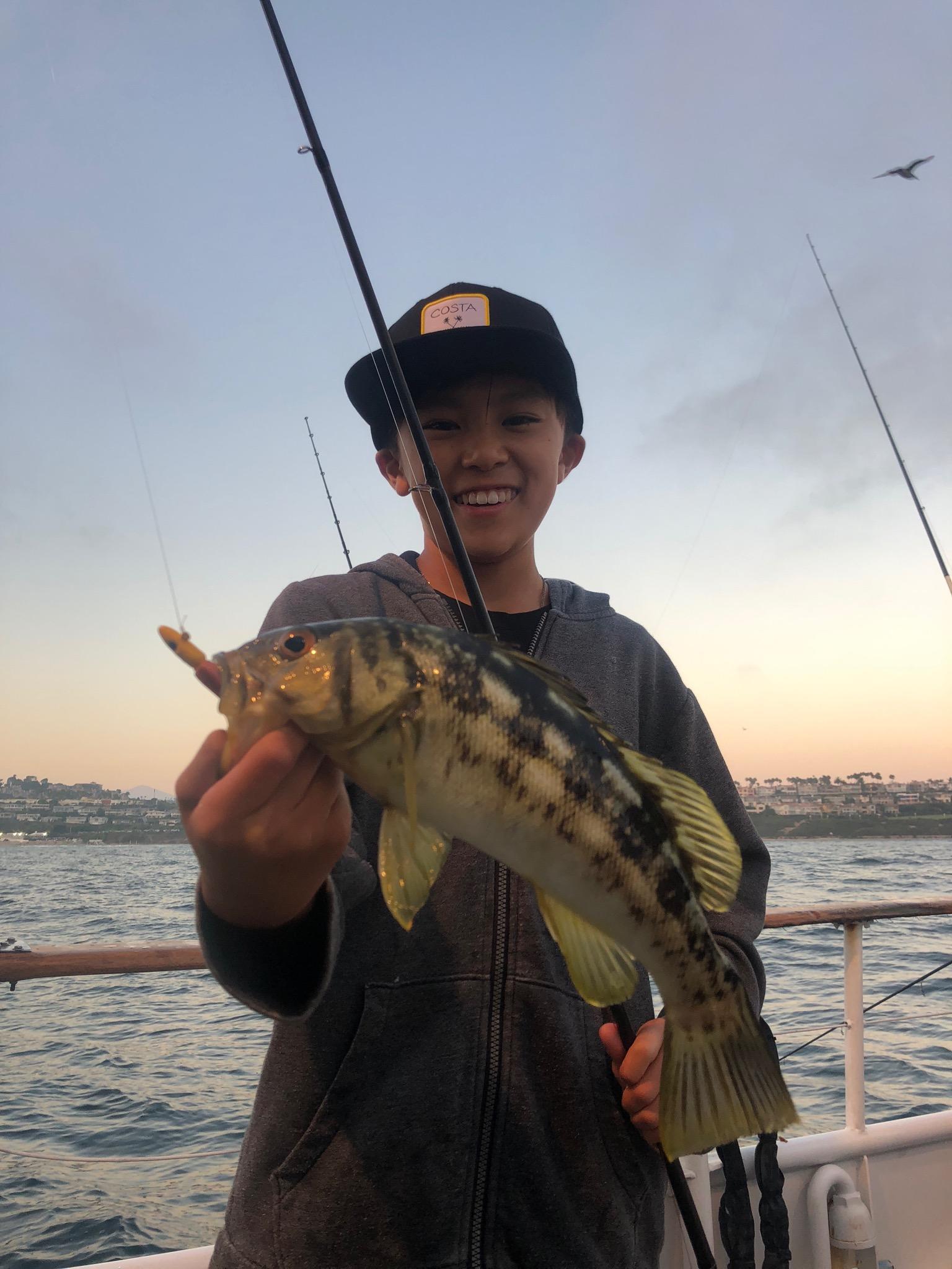 Smiling boy holding a fish on a boat with a sunset backdrop.
