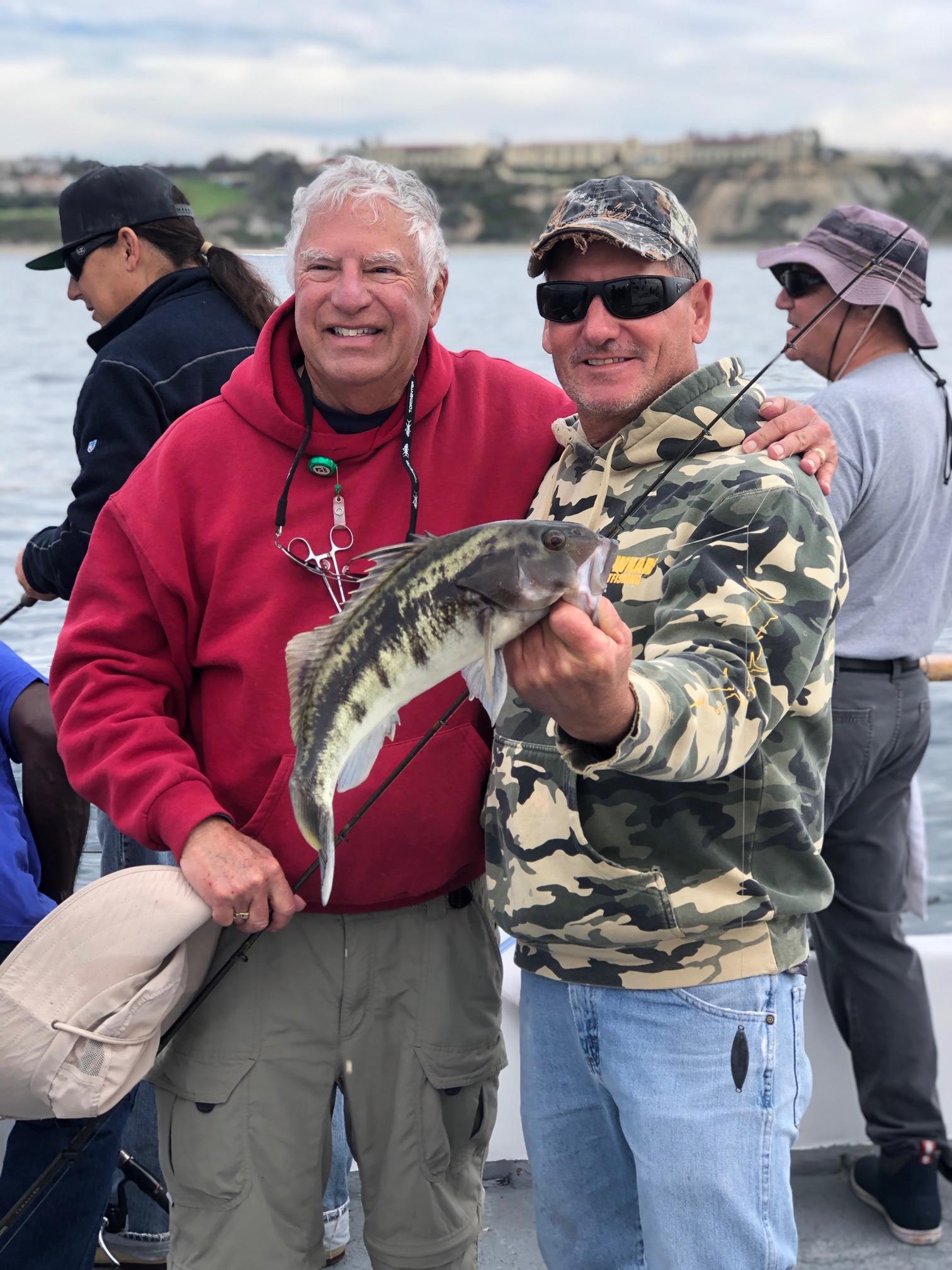 Two men smiling, one holding a fish on a boat with others in the background.