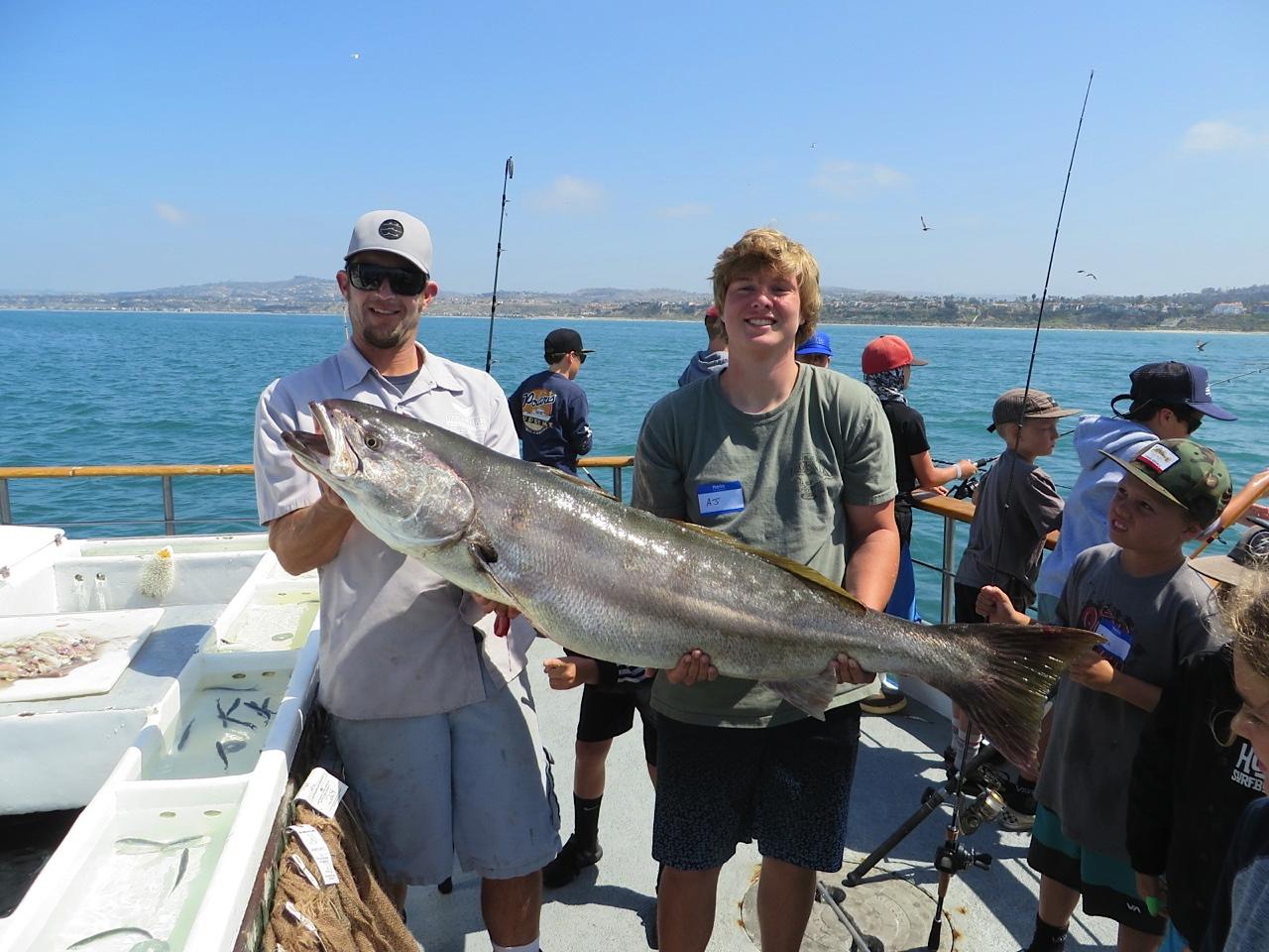 Two men holding a large fish on a boat, with others fishing in the background.