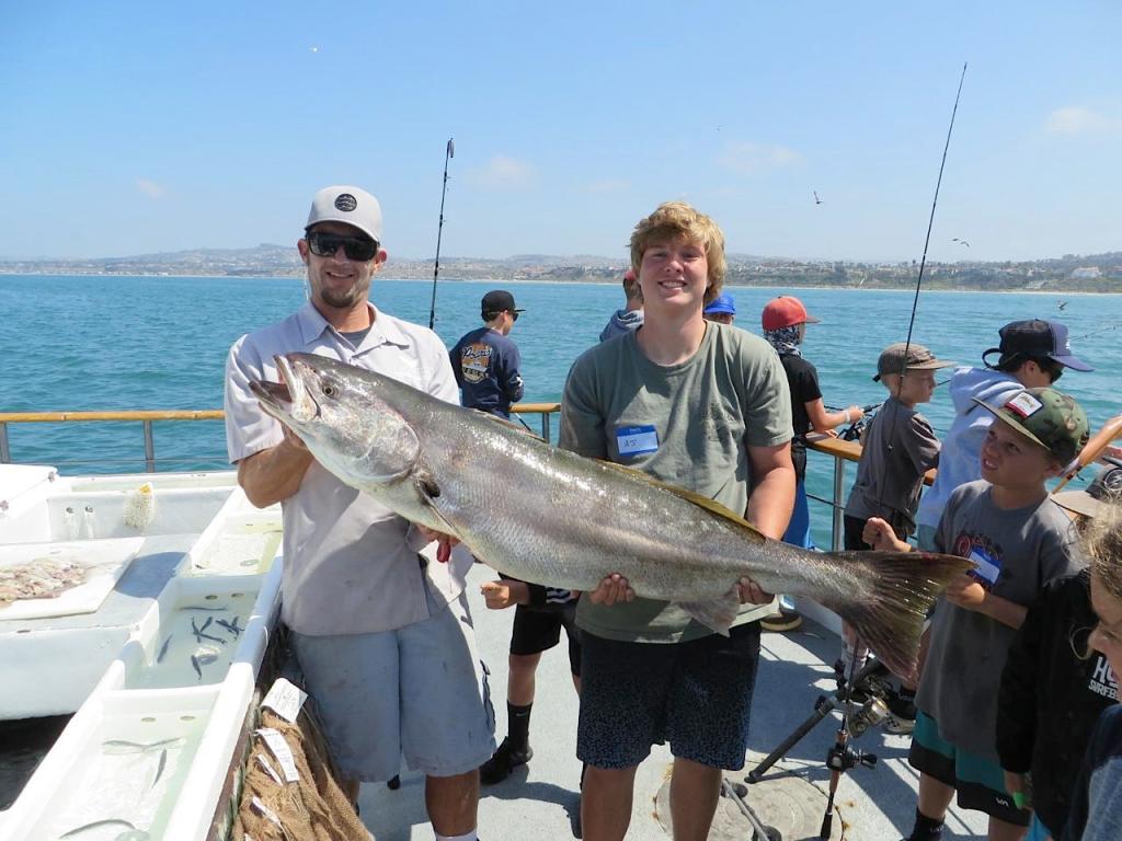 Two men holding a large fish on a boat, with others fishing in the background.
