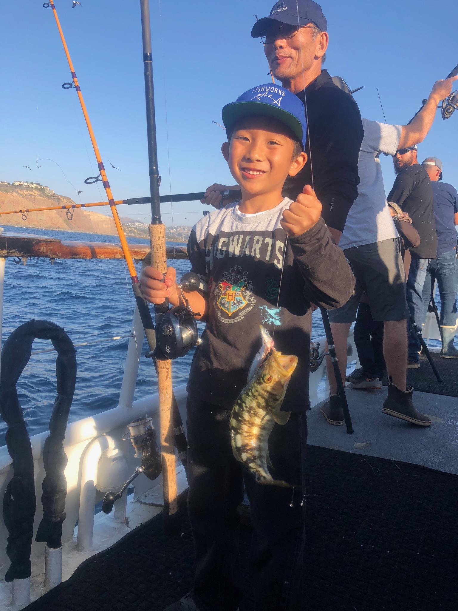 Child on a boat holding a fishing rod with a small fish, supported by an adult behind.