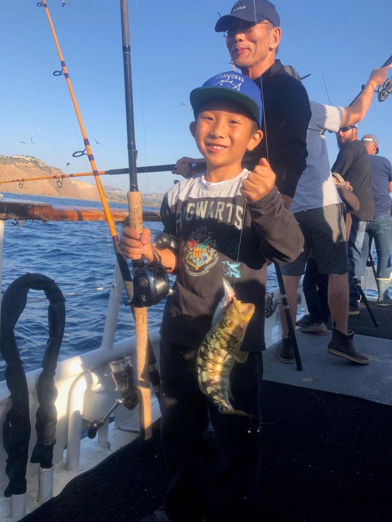 Child on a boat holding a fishing rod with a small fish, supported by an adult behind.