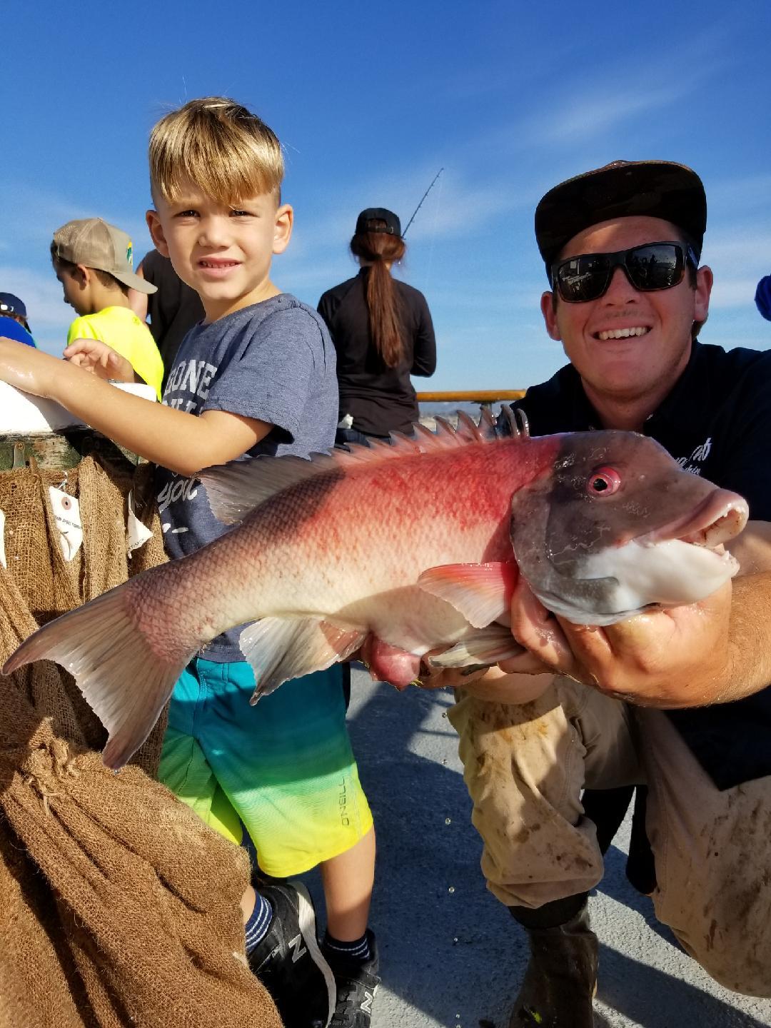 Man and boy on a boat holding a large fish under a clear blue sky.