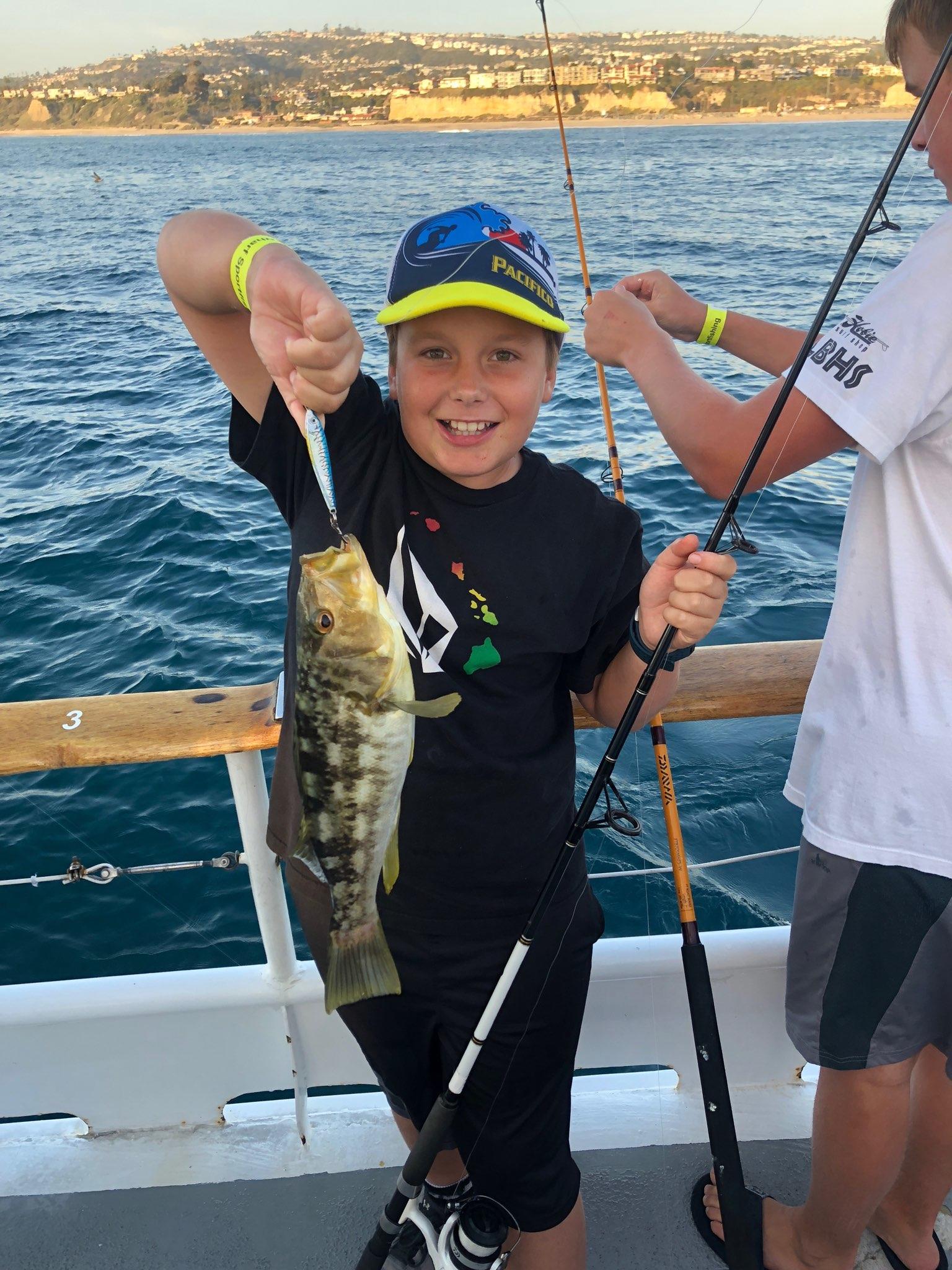 Boy smiling with a fish on a boat, ocean and cliffs in background.
