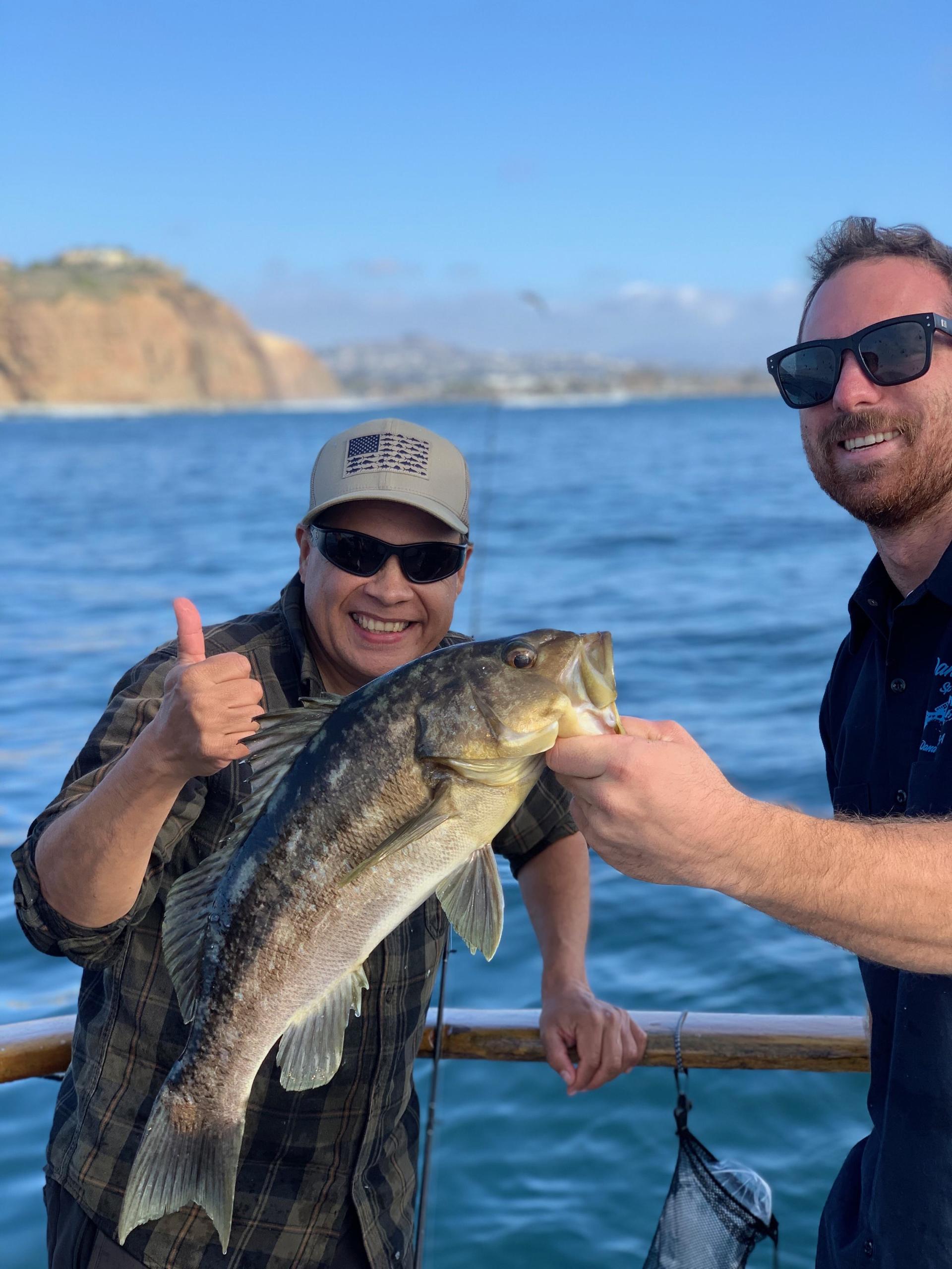 Two people on a boat holding a large fish, with ocean and cliffs in the background.