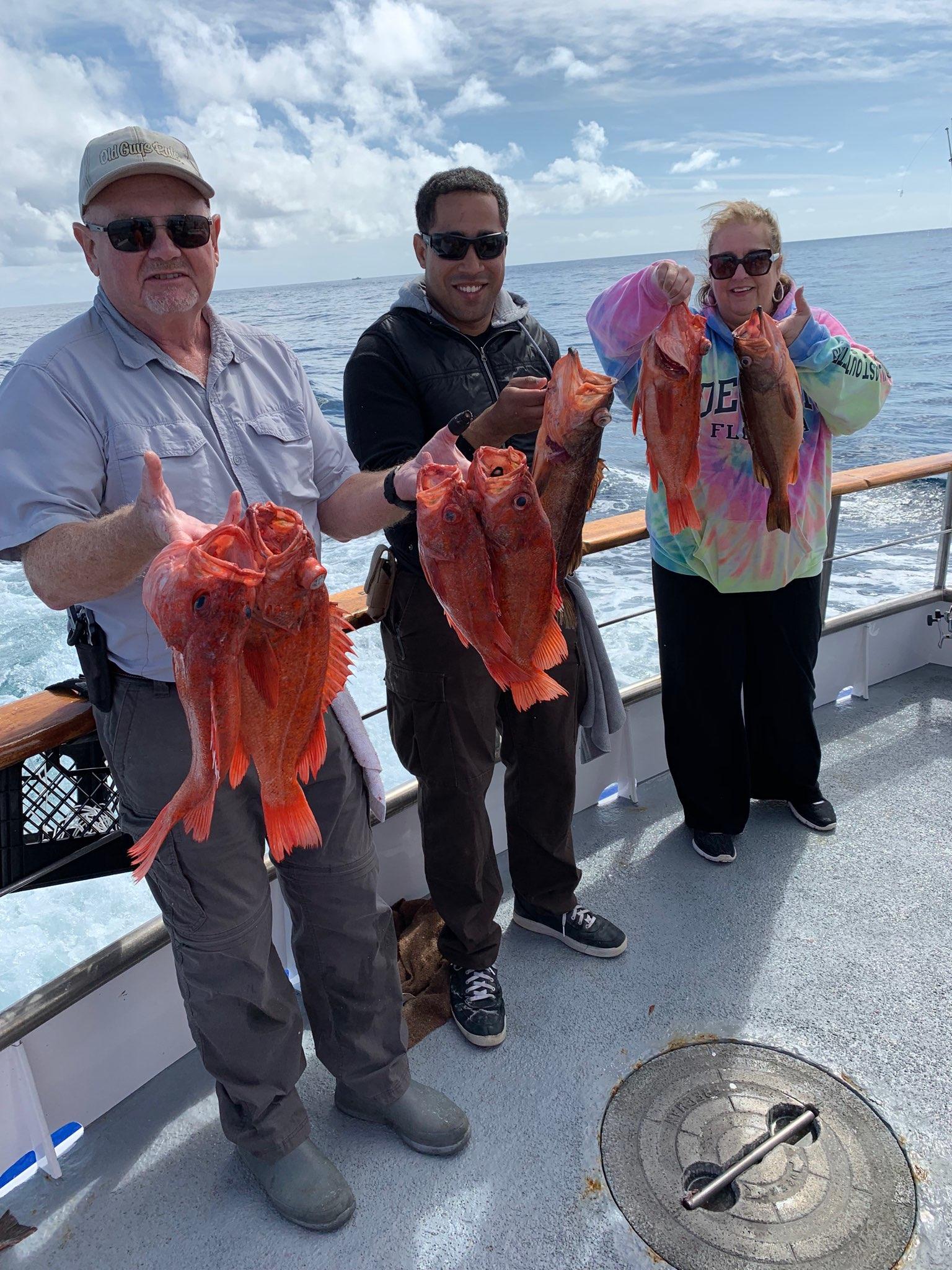 Three people on a boat holding several large red fish with the ocean in the background.
