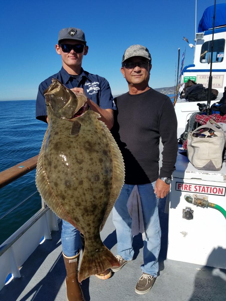 Two men on a boat holding a large fish, with clear blue sky in the background.