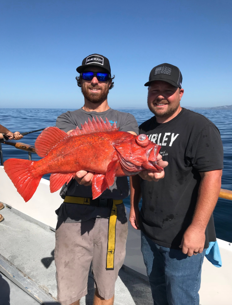 Two men smiling on a boat, holding a large red fish.