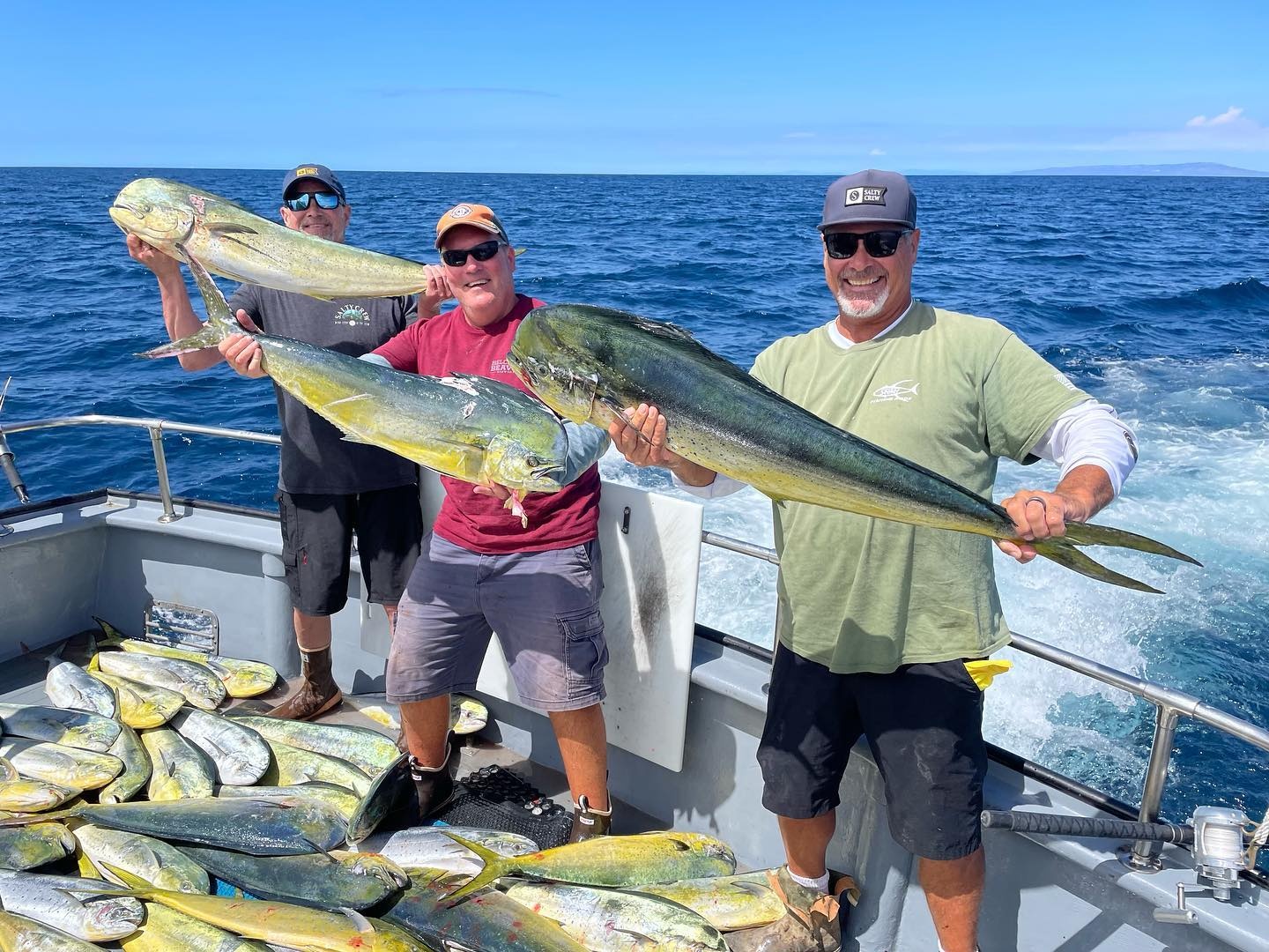 Three people on a boat holding large fish, with many fish at their feet.