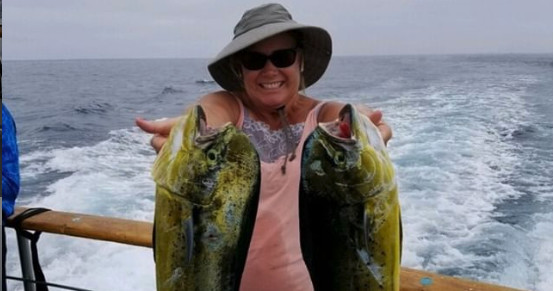 Smiling person holding two large fish on a boat at sea.