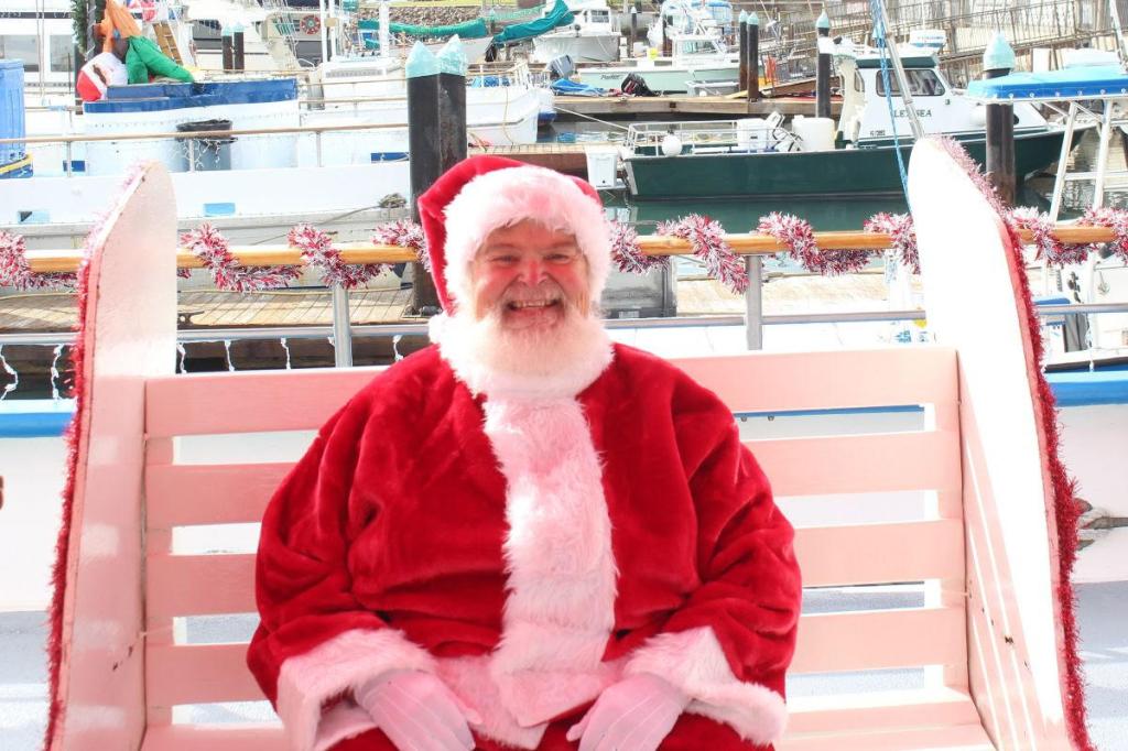 Santa Claus sitting on a bench with boats in the background.