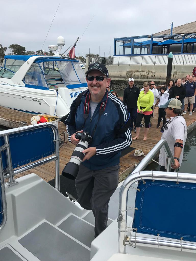 Man with camera on boat, people in background on dock.