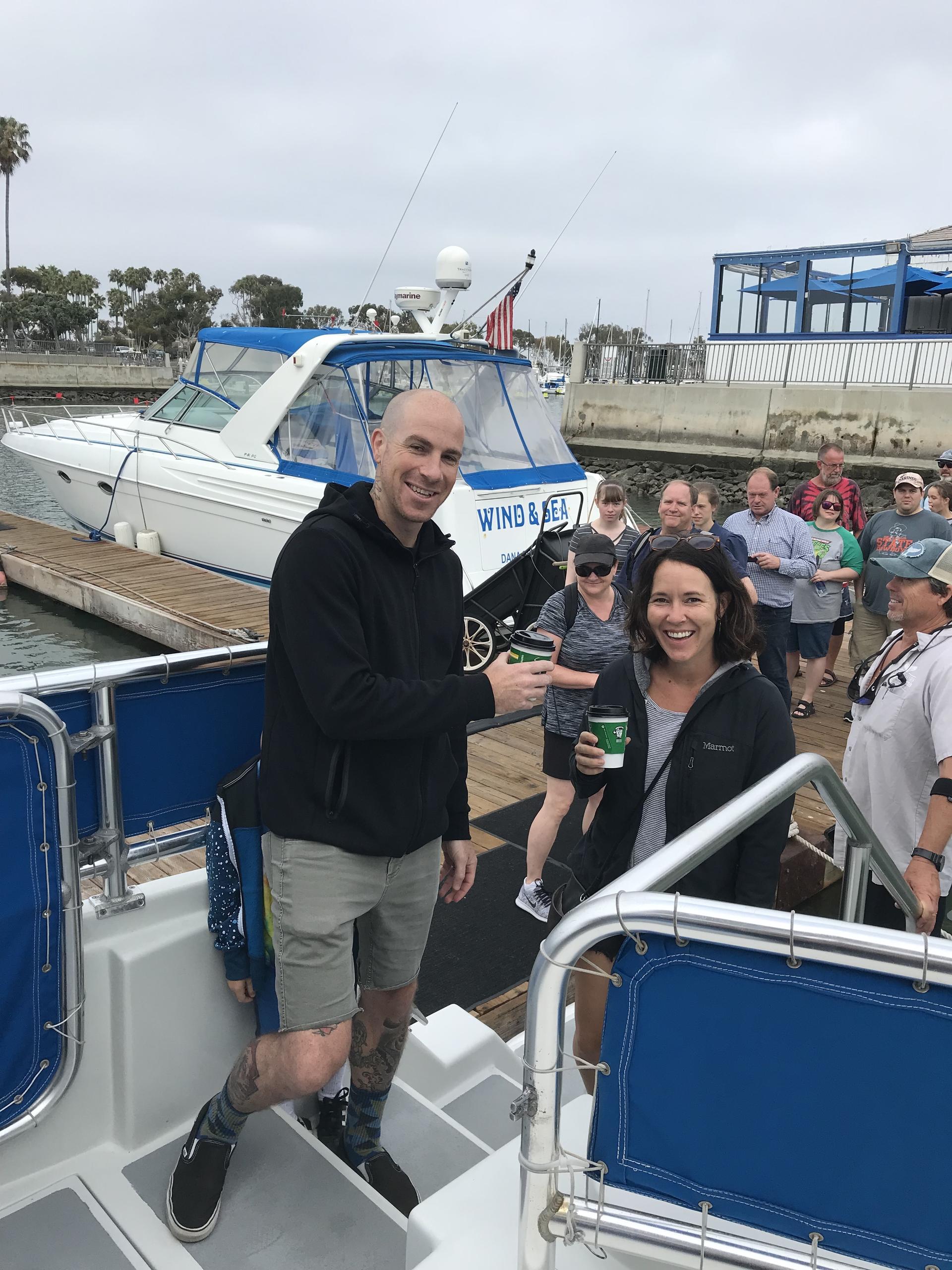 Two people smiling on a boat with others and a marina in the background.