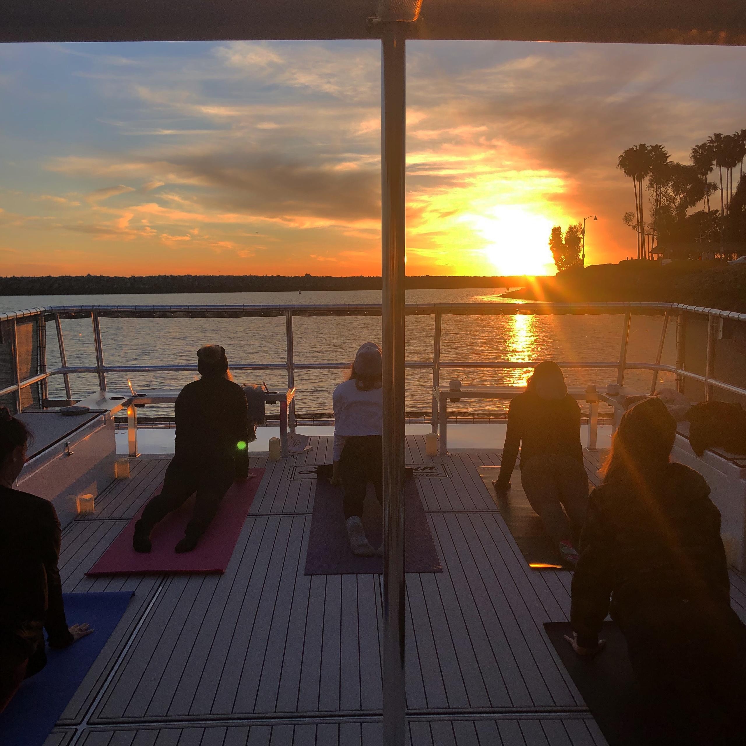People doing yoga on a boat deck at sunset, facing the water.
