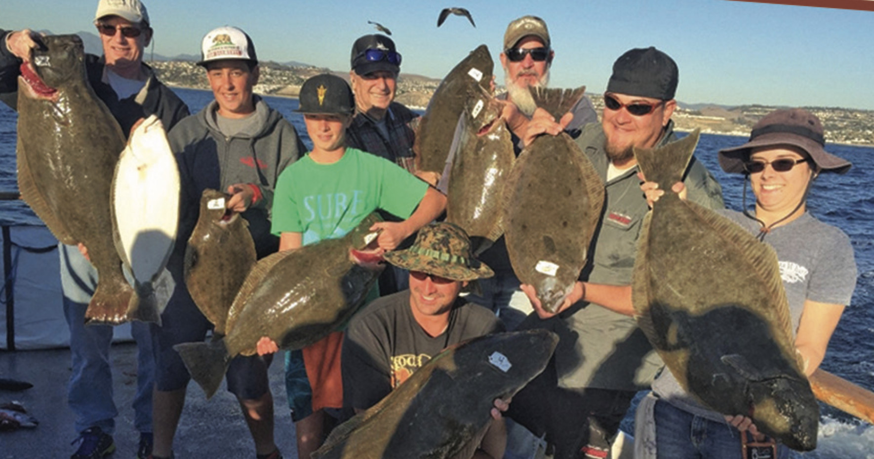 Group of people on a boat holding large fish, with ocean and coastline in the background.