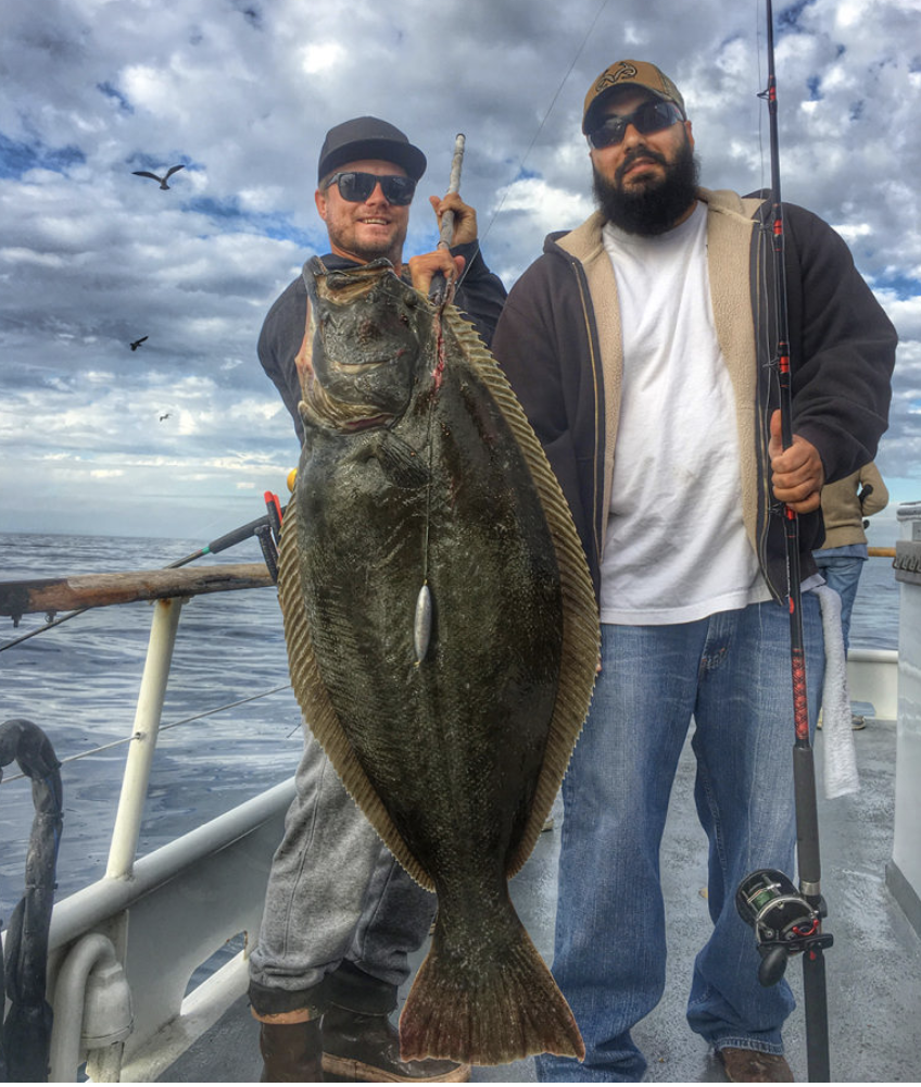 Two men on a boat holding a large fish, with a cloudy sky and ocean in the background.