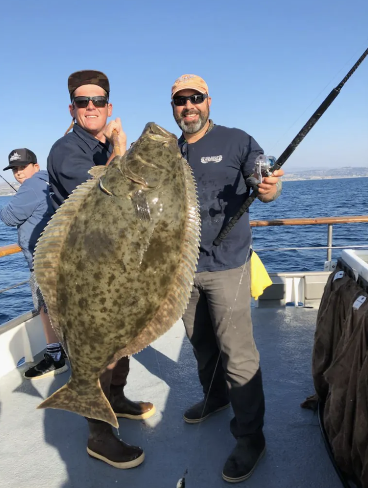 Two men on a boat holding a large flat fish and a fishing rod.