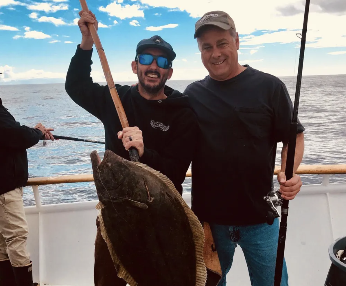 Two men smiling with a large flatfish on a fishing boat.