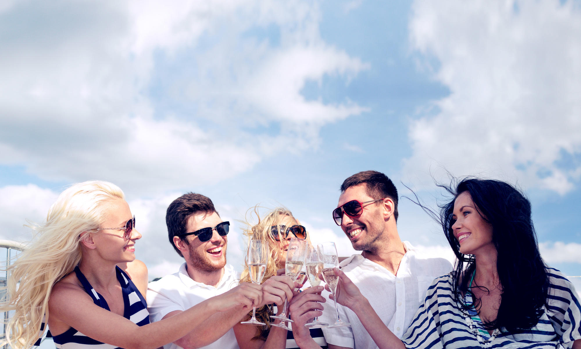 Five people outdoors clinking glasses under a blue sky with clouds.
