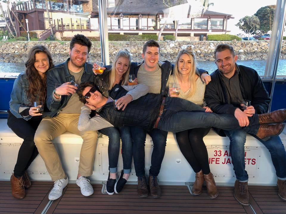 Group of seven friends on a boat, one person lying across laps, all smiling and holding drinks.