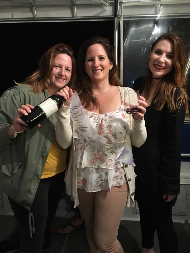 Three women smiling indoors, one holding a bottle, another wearing a 'Bride to Be' sash.