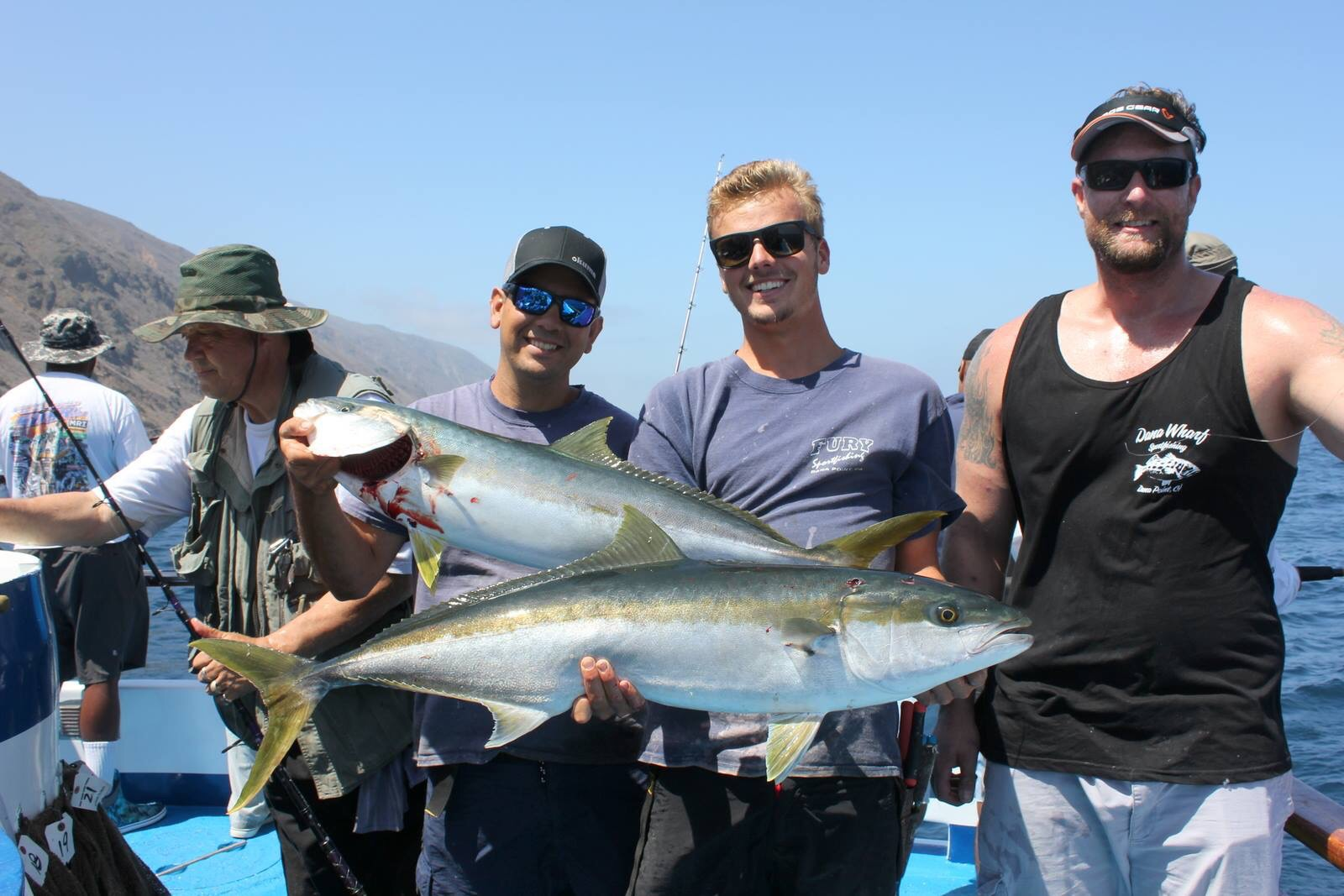 Four people on a boat holding two large fish, with mountains and sea in the background.