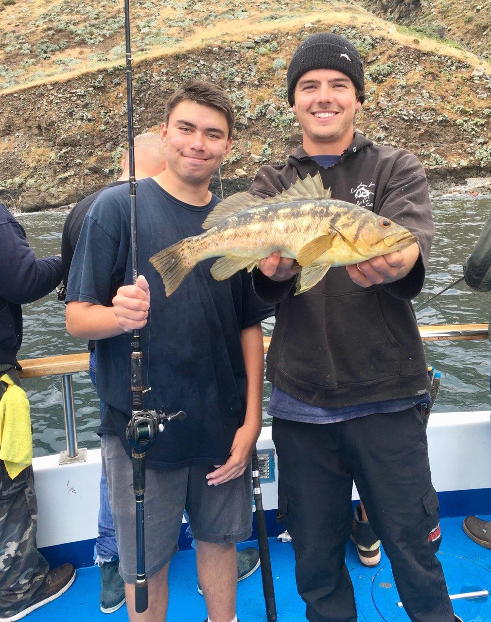 Two people on a boat, one holds a fish and a fishing rod, both smiling.
