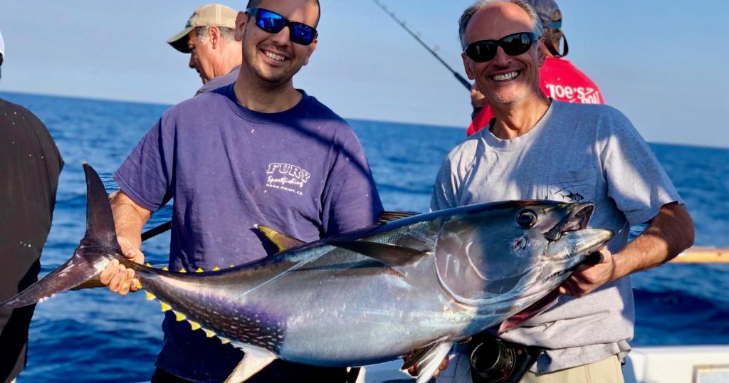 Two smiling men holding a large fish on a boat at sea.