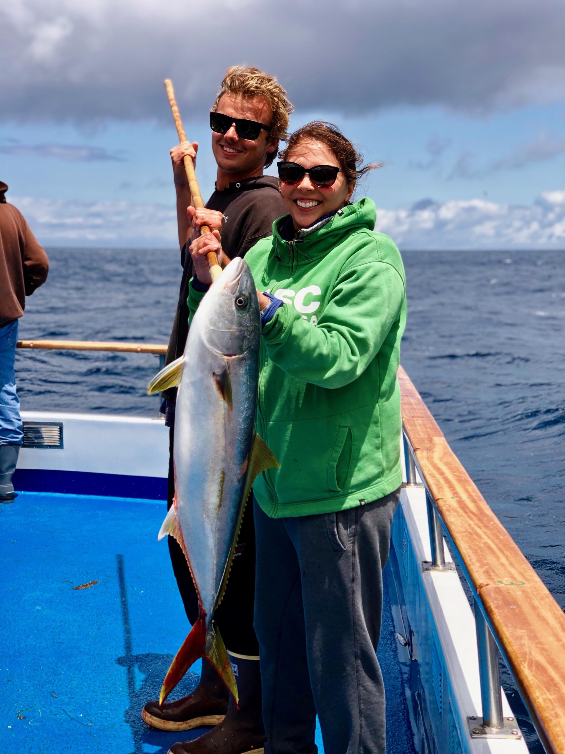 Two people on a boat holding a large fish, smiling, under cloudy skies.