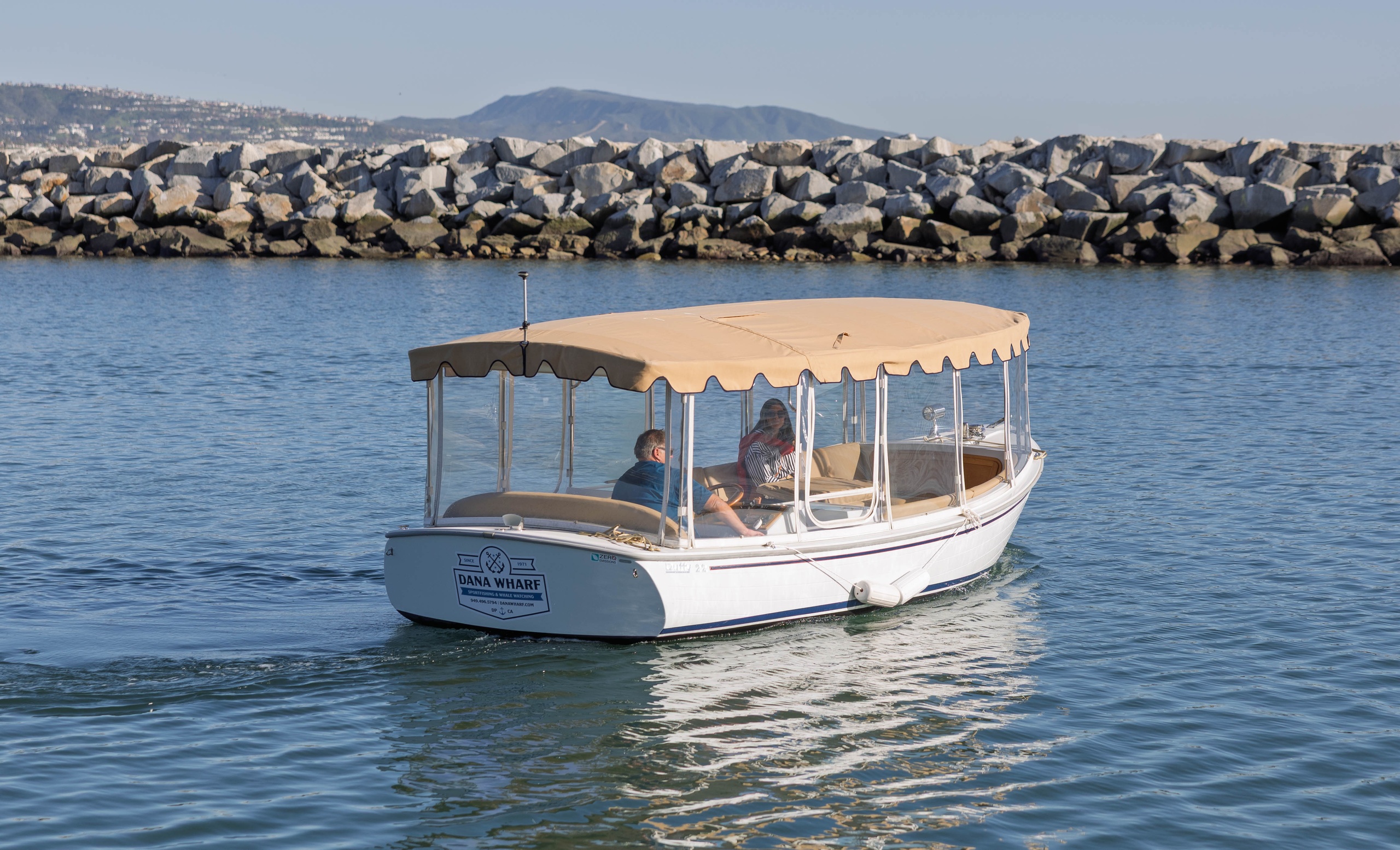 Small boat with canopy on calm water near rocky shoreline.