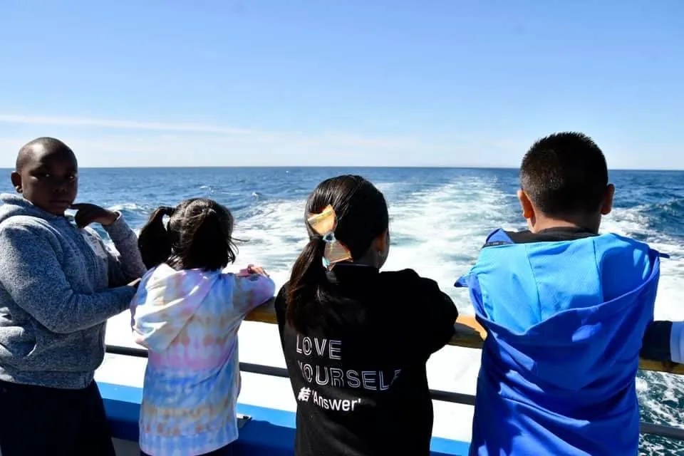Four kids on a boat looking at the ocean under a clear sky.