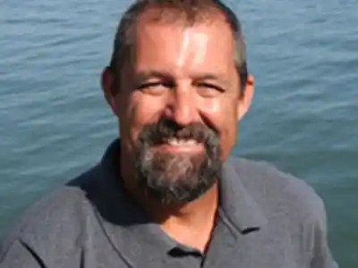 Smiling man with short hair and beard, wearing a gray shirt by water.