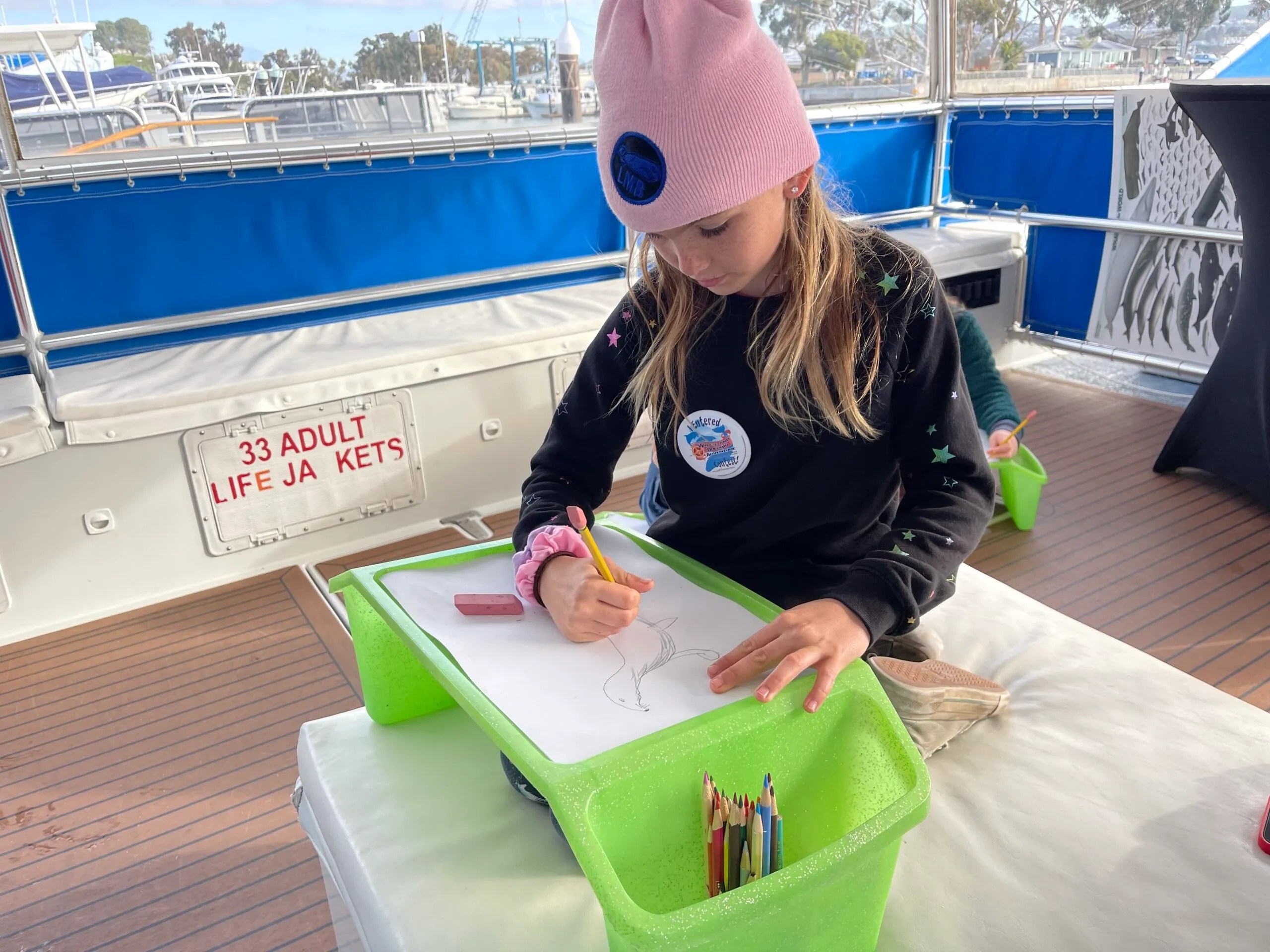 Child drawing on a boat with colored pencils, wearing a pink beanie.