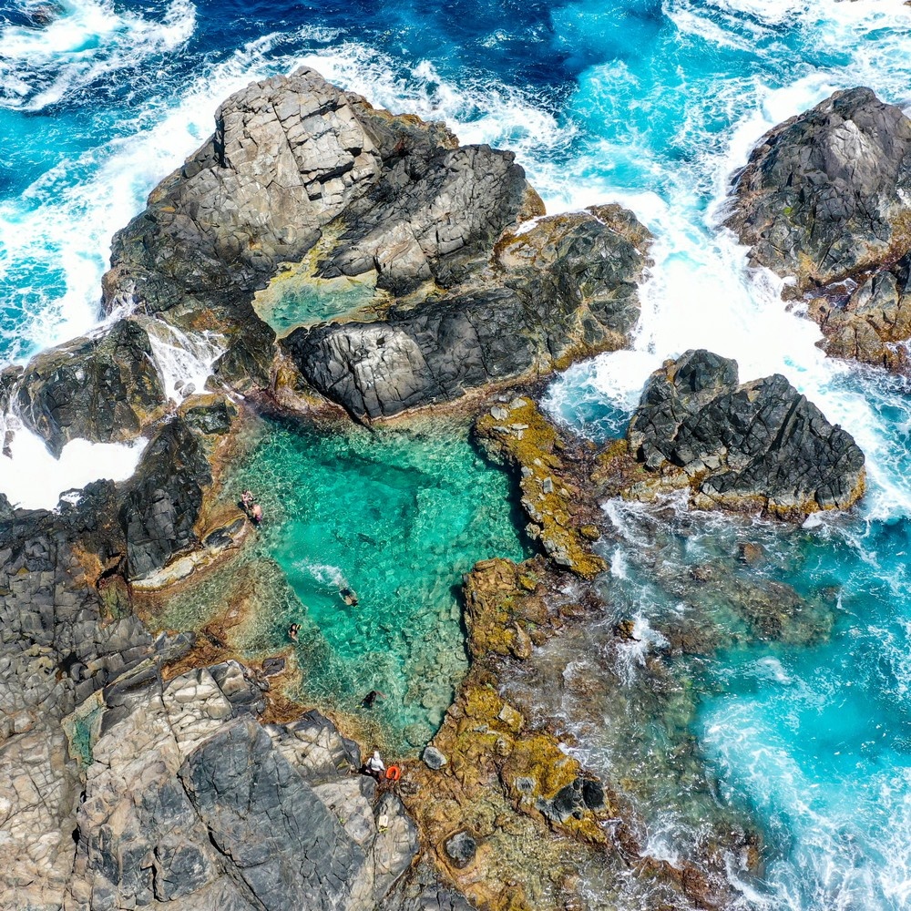 Aerial view of rocky coastline with turquoise tidal pool and crashing waves.