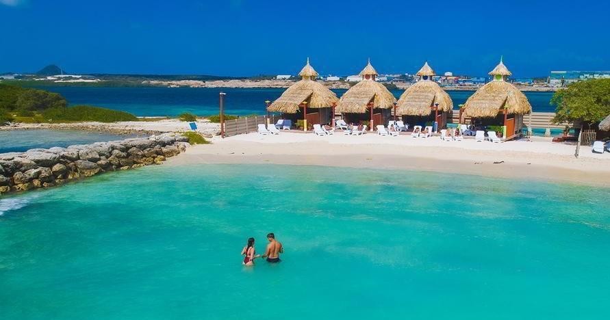 Two people wade in turquoise water near straw huts on a beach.