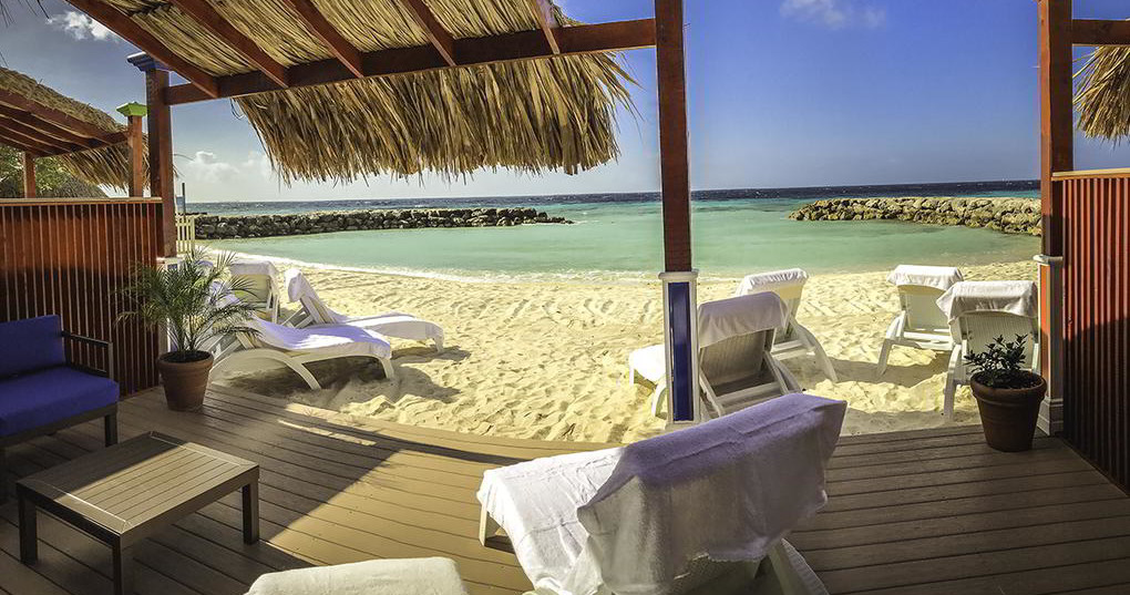 Beach view with lounge chairs, thatched roof, and clear blue water.