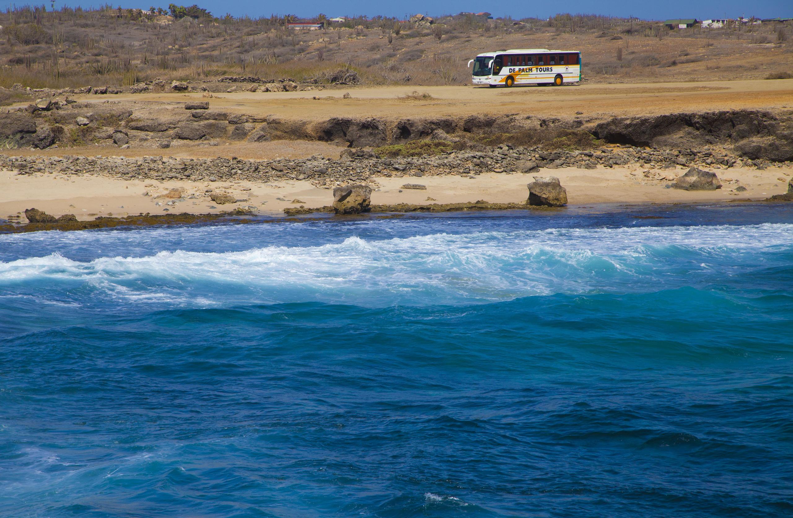 Tour bus on dirt road near rocky beach with ocean waves.