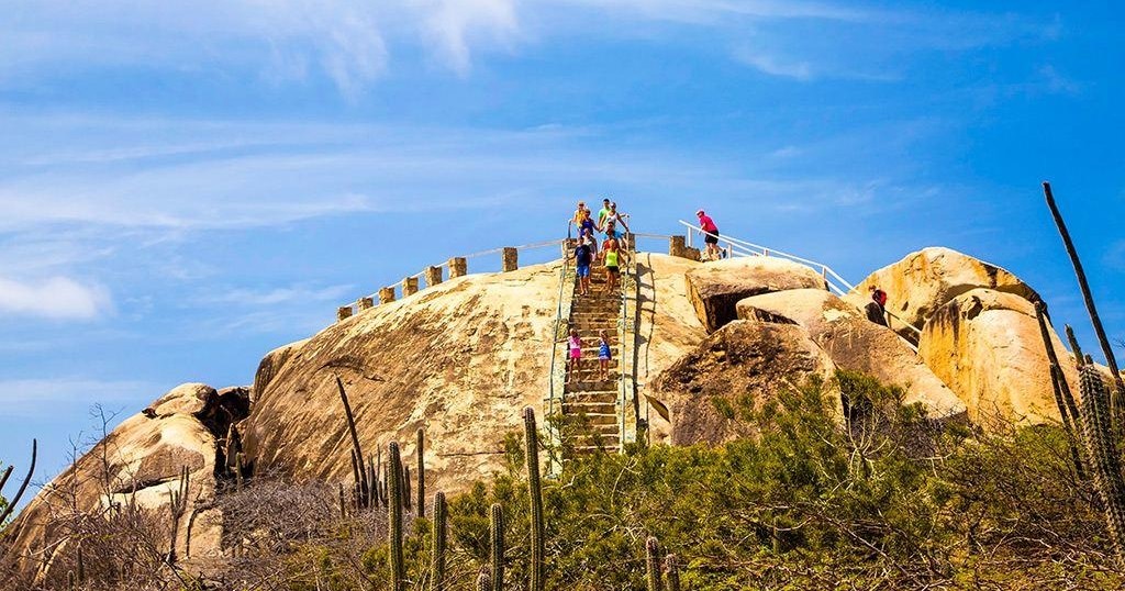 People climbing stairs on a large rocky hill under a blue sky.