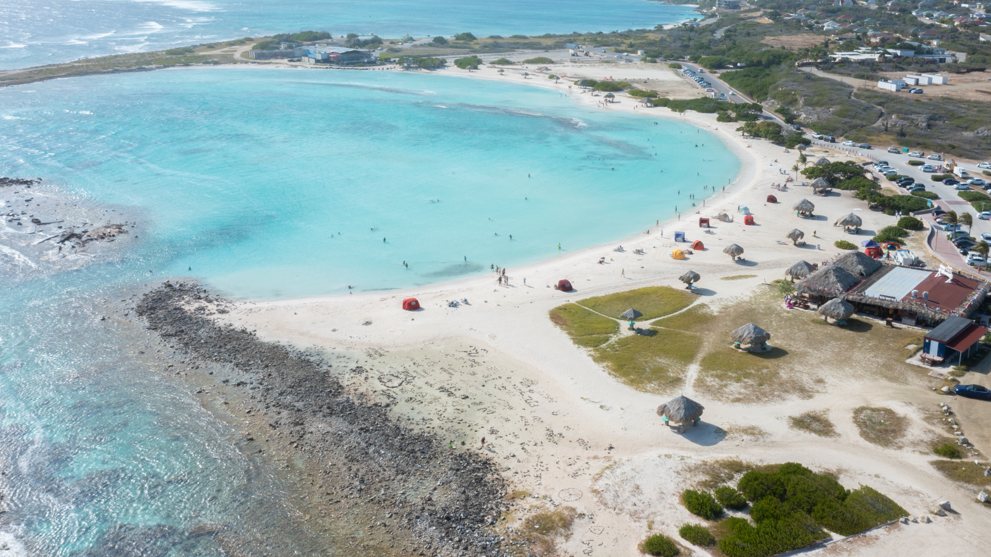 Aerial view of a beach with clear water, scattered umbrellas, and people swimming.