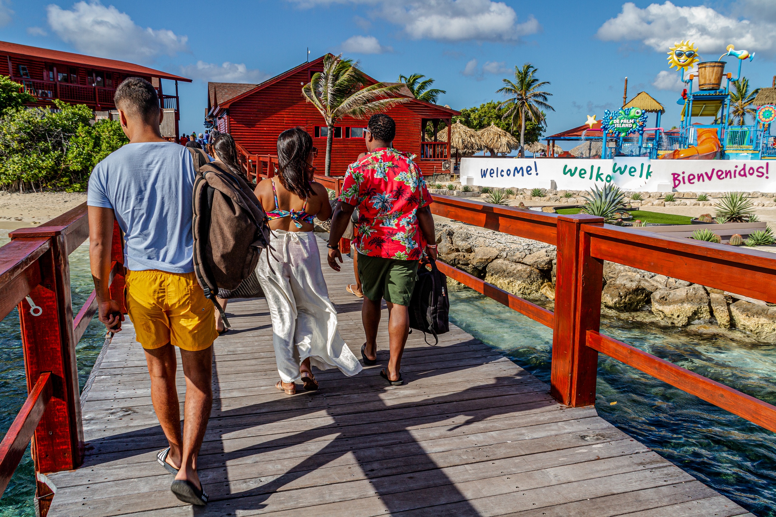 Three people walking on a wooden boardwalk toward colorful buildings and a welcome sign.