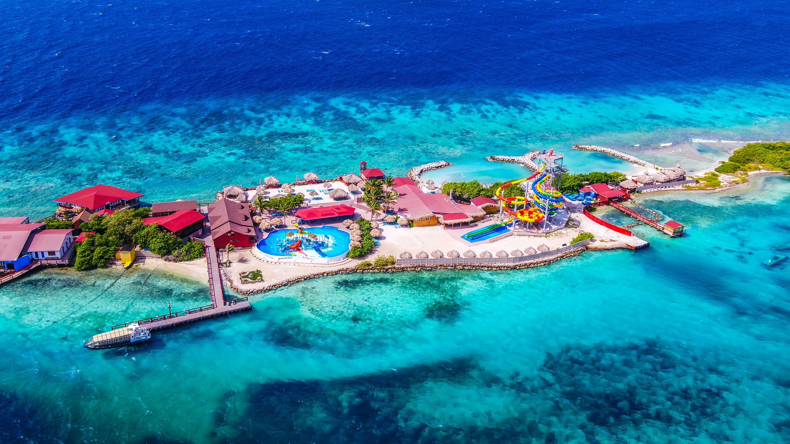 Aerial view of a colorful island resort with pools and slides, surrounded by turquoise ocean water.