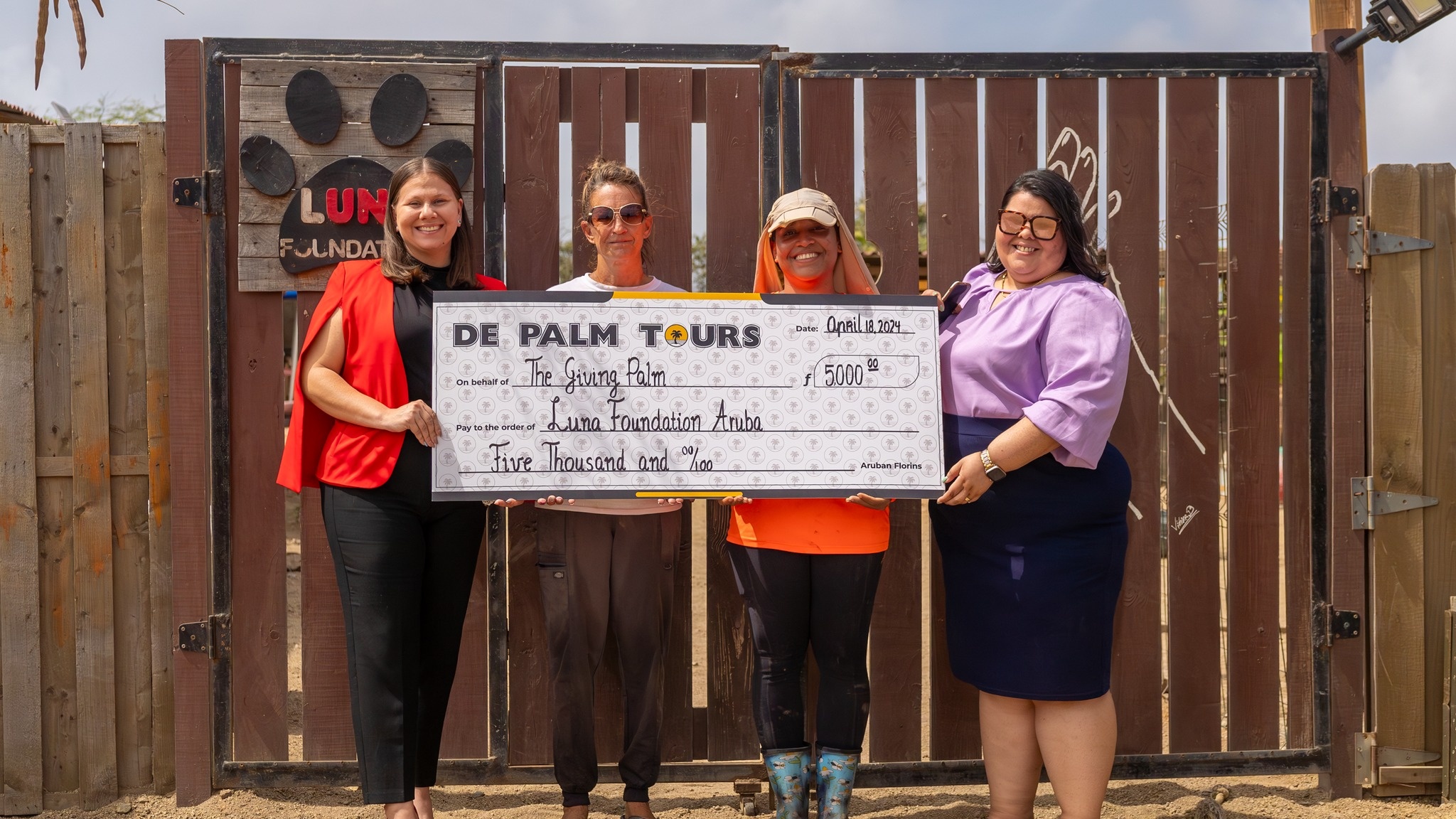 Four people hold a large donation check in front of a wooden gate.