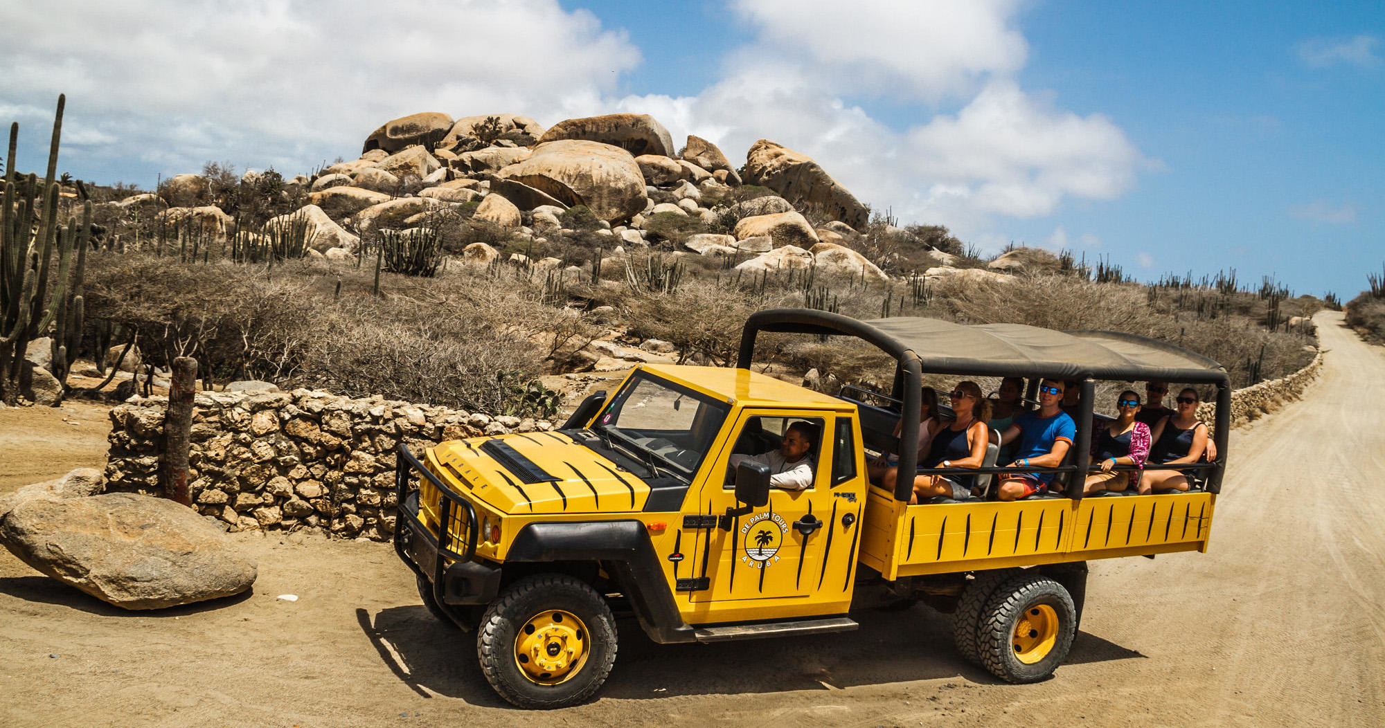 Yellow open-top safari vehicle on dirt road with passengers, rocky desert landscape.