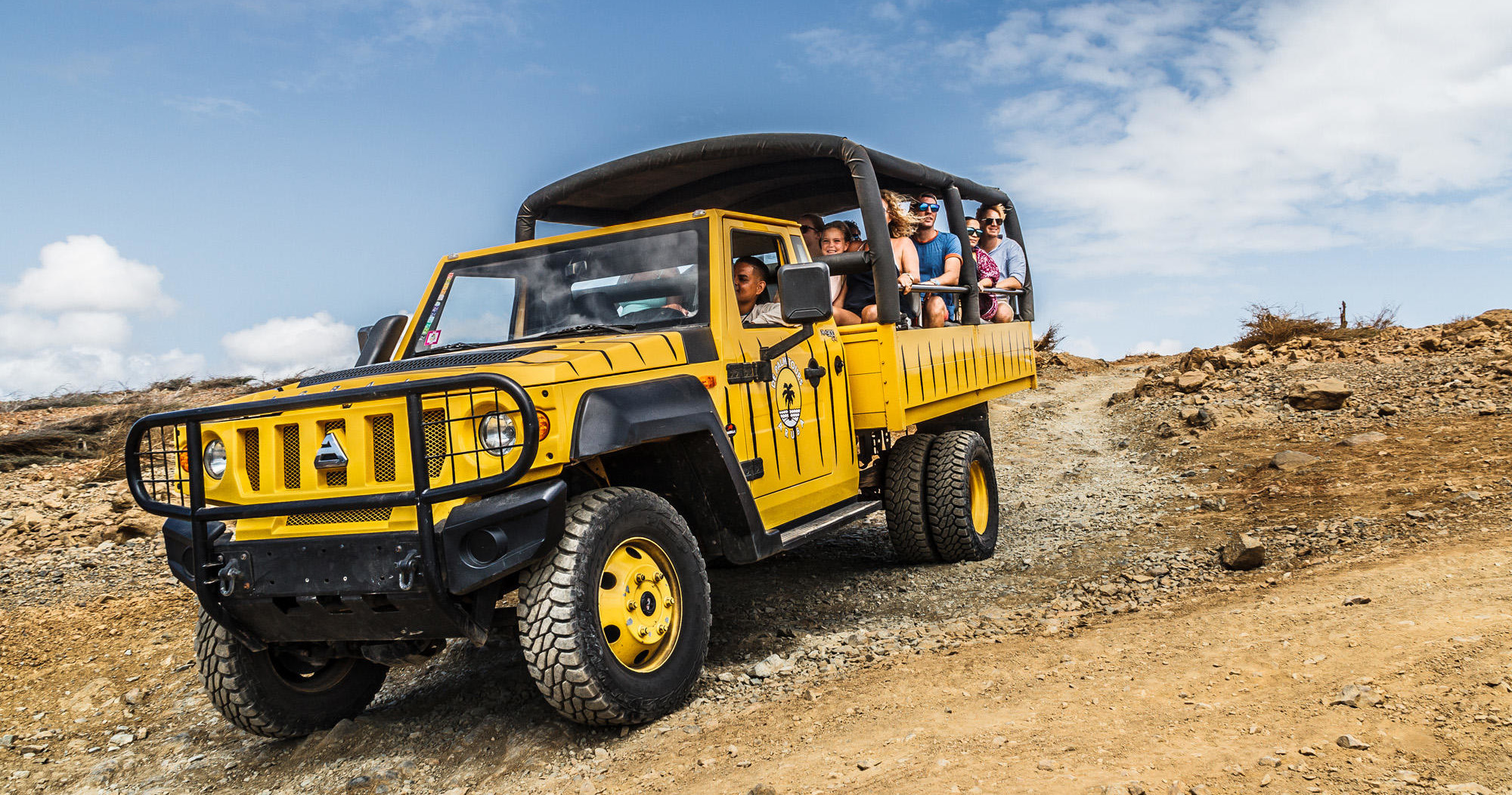 Yellow safari vehicle with tourists on rocky terrain under blue sky.