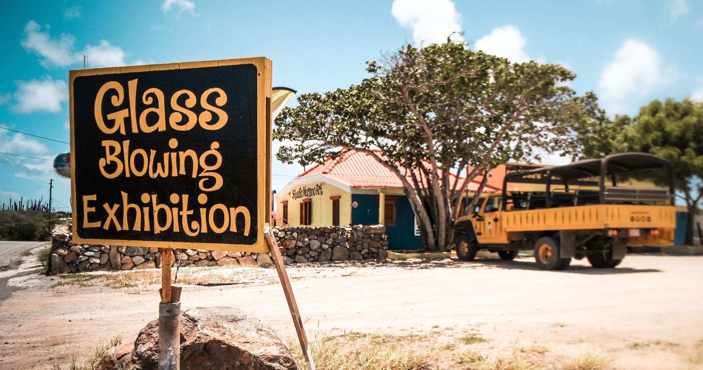 Sign for Glass Blowing Exhibition near a rustic building and yellow truck.