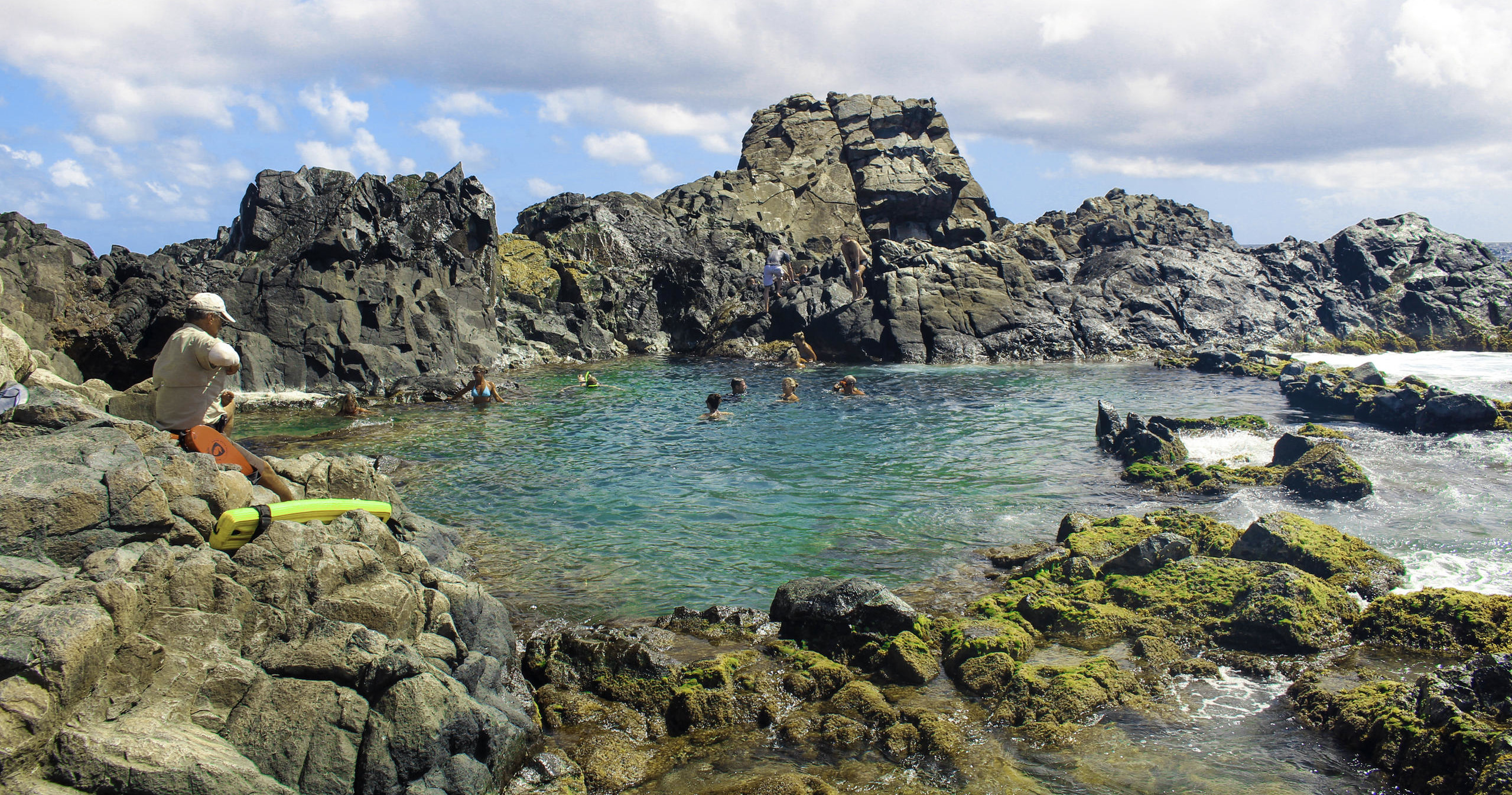 Natural rock pool with people swimming, surrounded by jagged rocky formations.